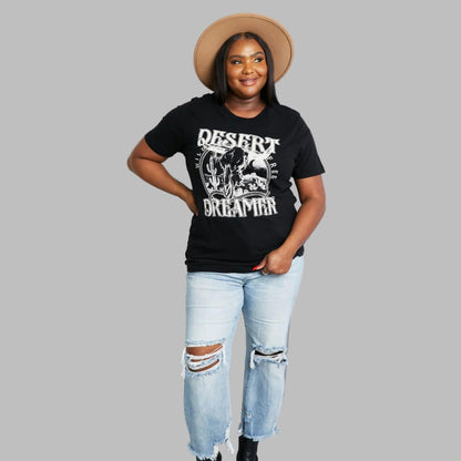 Person wearing a black t-shirt with 'Desert Dreamer' text and graphics, light blue ripped jeans, and a beige hat.
